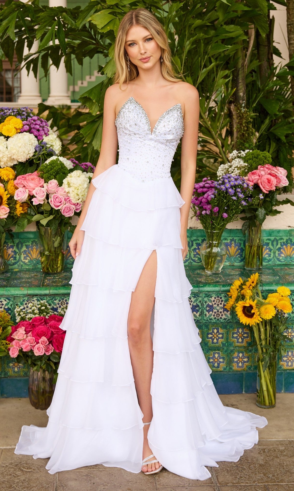 A woman in the Ava Presley 26407 strapless ruffled prom gown with a chiffon a-line skirt and thigh-high slit stands before a flower display and green foliage.